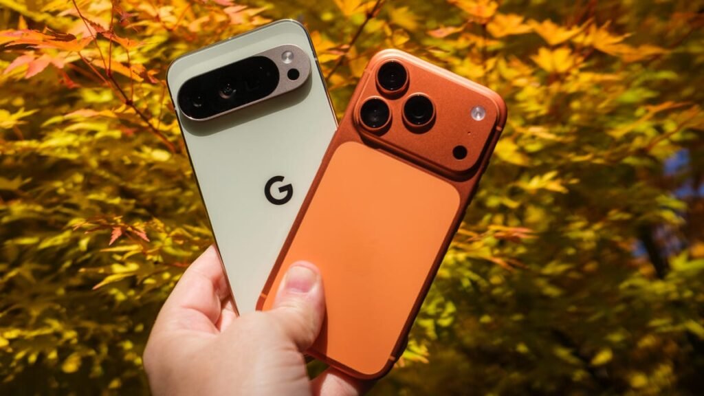 Hand holding a light green Google Pixel 10 Pro XL and an orange Apple iPhone 17 Pro in front of a tree with orange and green leaves.